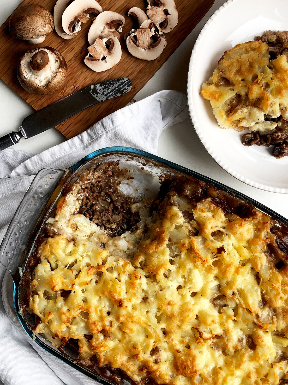 Golden brown cottage pie in a Pyrex oven dish and sliced mushrooms on a chopping board