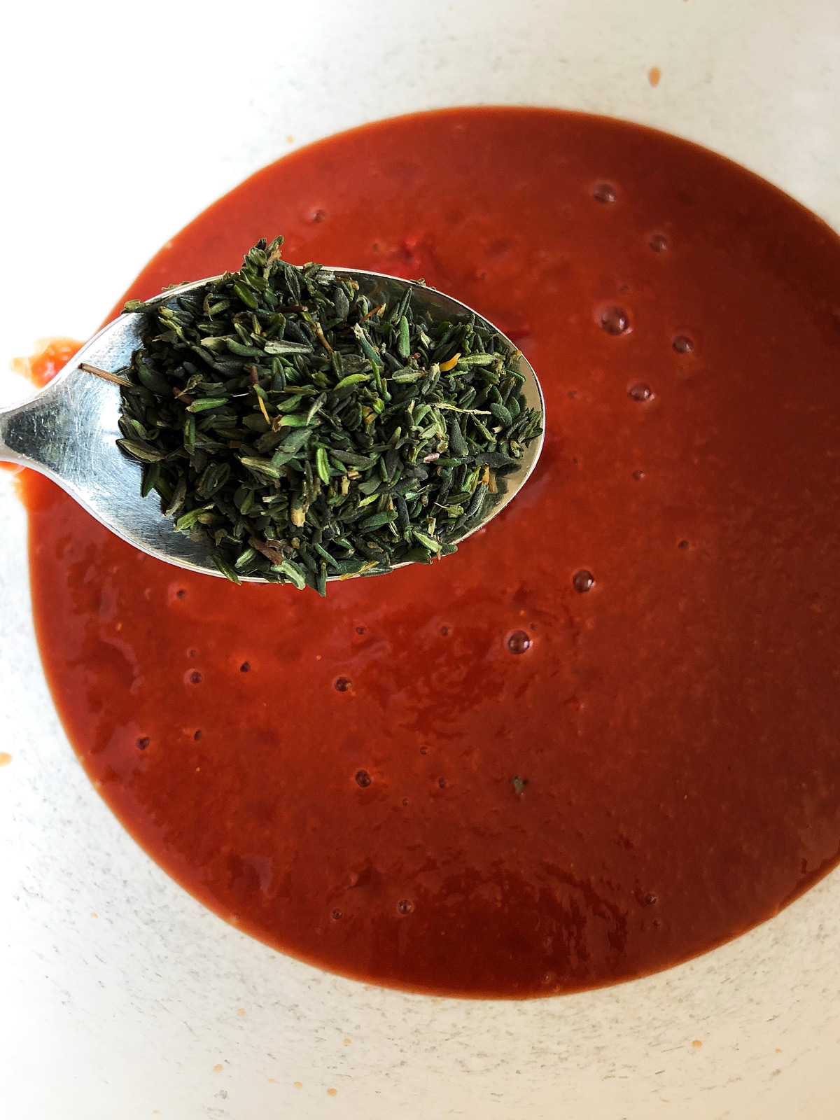 Fresh thyme leaves on a spoon with red passata in the background.