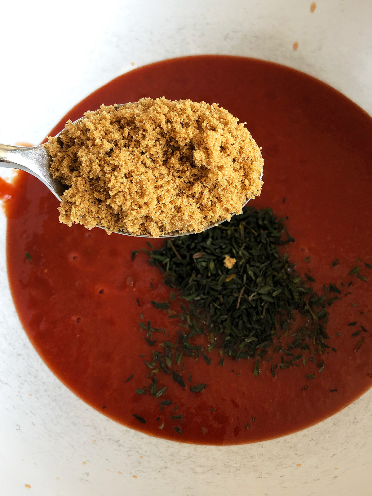 A spoonful of muscovado sugar above a bowl with passata and herbs.