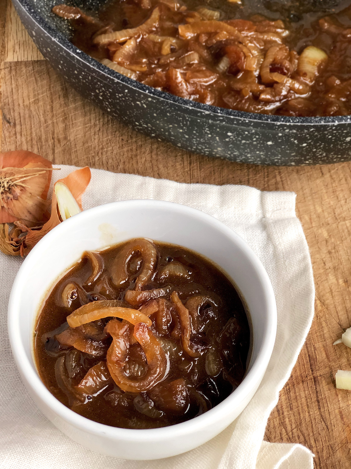 Close up - onion gravy in a small white bowl with a non-stick pan with more gravy at the background.