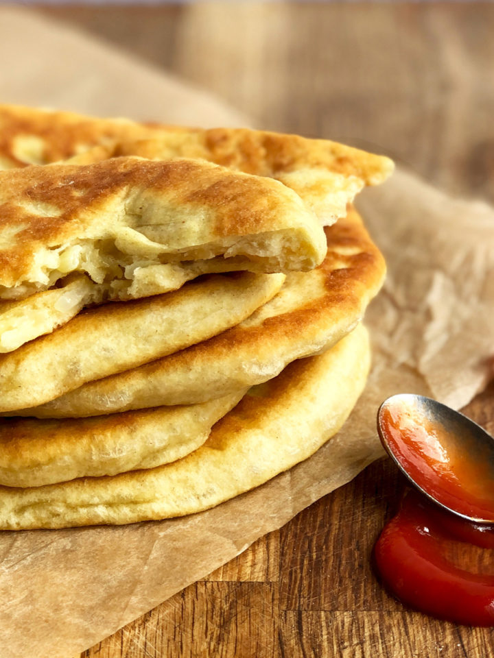 A pile of golden brown fried flatbreads on a wooden chopping board with a teaspoon of ketchup next to them