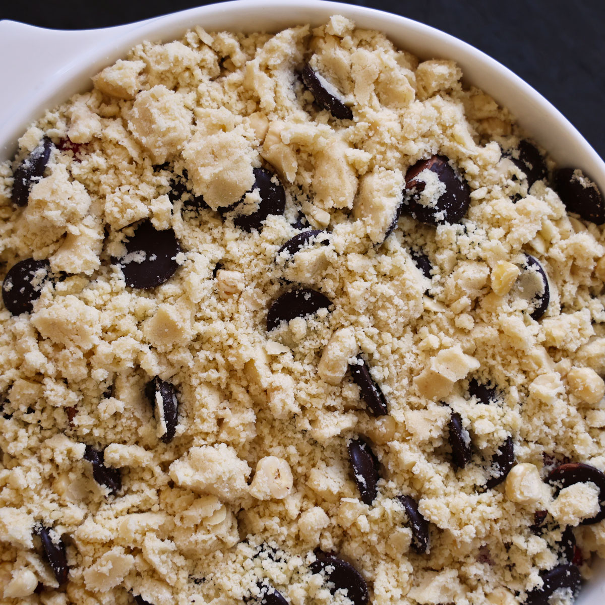 Asembled crumble with lumpy topping with pieces of chocolate and crushed hazelnuts evenly spread over the fruit.