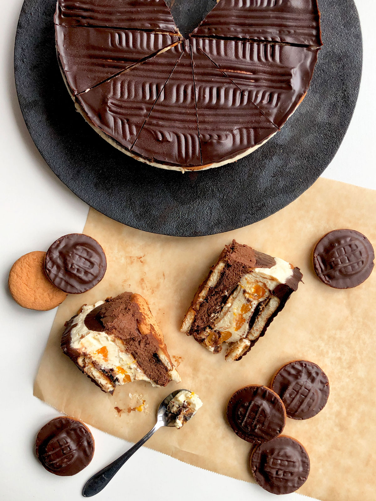 Bird-eye view: two slices of a whipped cream cheesecake, round chocolate covered cake with a few Jaffa cakes scattered around. A round Jaffa cake cheesecake on a cast iron