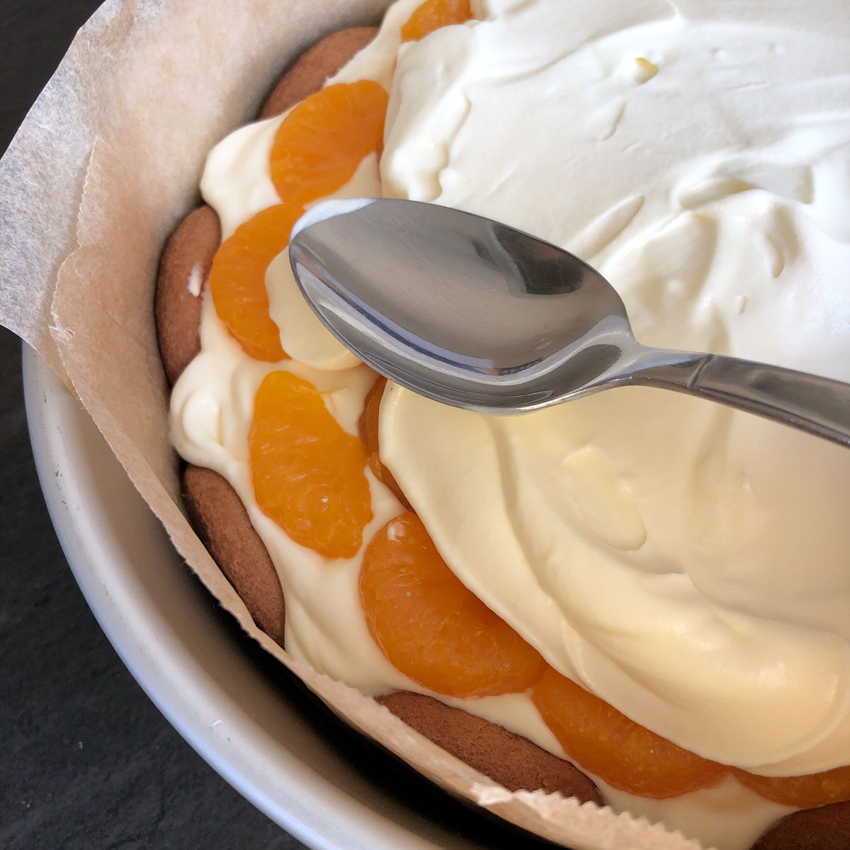 A spoon spreadding whipped cream over a layer of tinned mandarins.