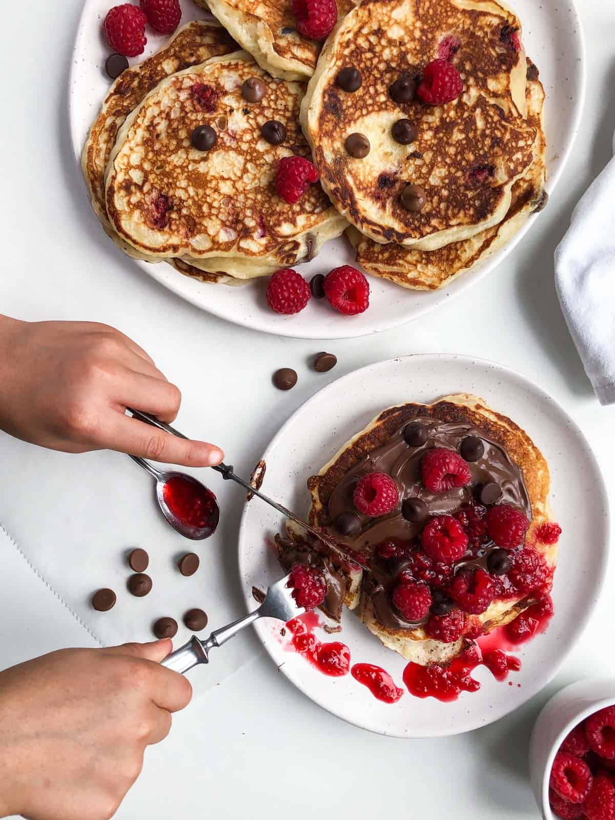 Hands holding cuttlery, cutting pancake topped with chocolate, raspberries and chocolate chips.