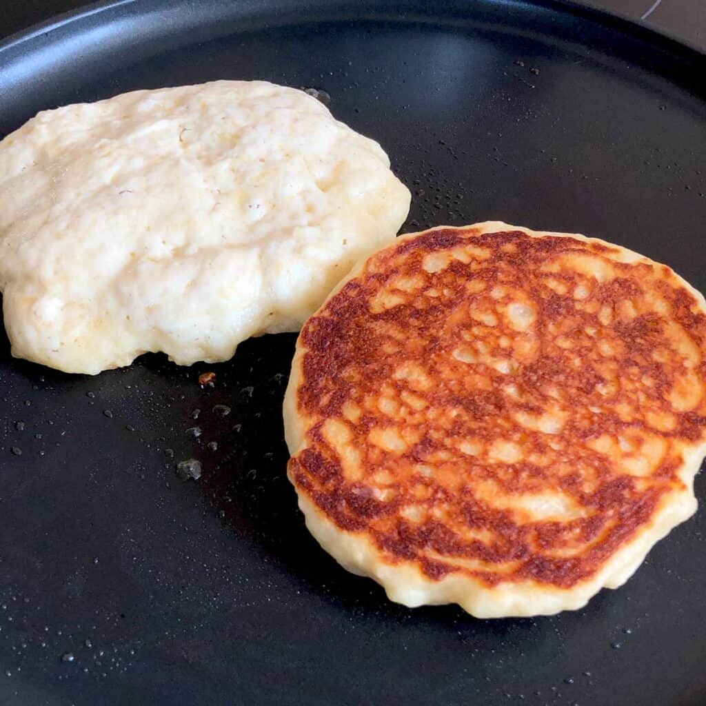Two pancakes in the hot pan; one flipped over with golden-brown top.